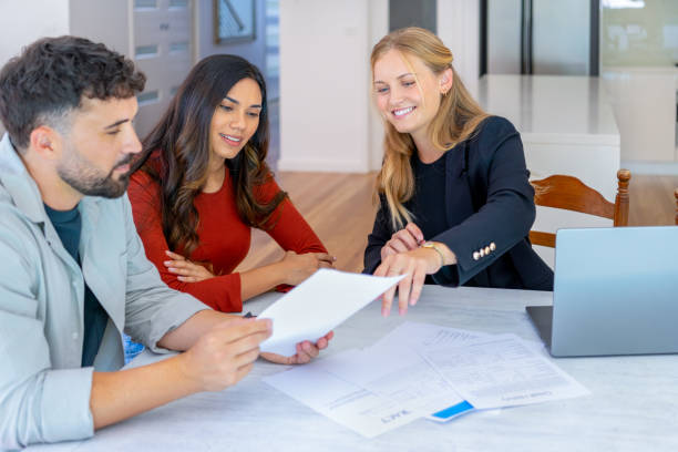 Real Estate or Insurance agent with couple looking through documents. Couple are casually dressed. They sitting at a table at home and are looking a little happy and smiling. There is a laptop computer on the table. The kitchen and living room can be seen in the background