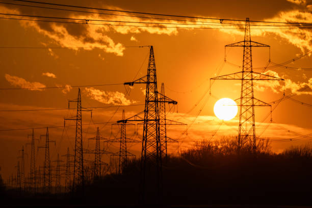 The setting sun casts a warm glow behind silhouetted powerlines, highlighting the infrastructure's intricate design as it stretches across the landscape.