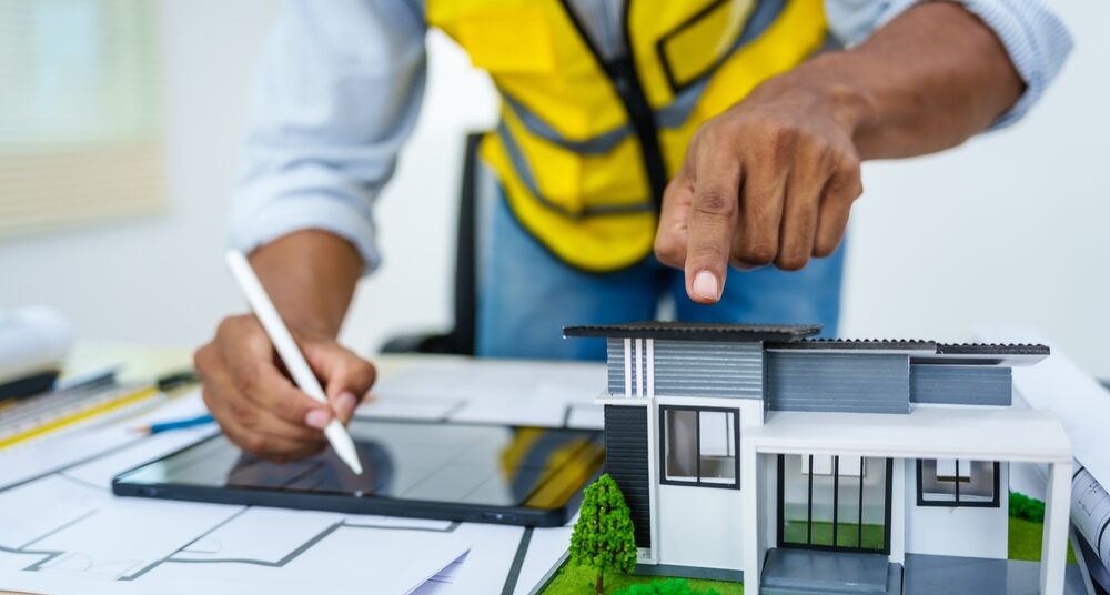 Asian male architect works at desk with house model toy and A3 paper blueprints, construction real estate law, zoning, permits, Los Angeles Zoning Codes