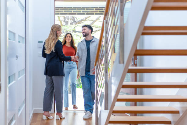 Real estate agent showing a young couple a new house. The home is contemporary. All are happy and smiling. The couple are casually dressed and the agent is in a suit and shaking hands .The couple are being greeted at the front door by the real estate agent. Copy space