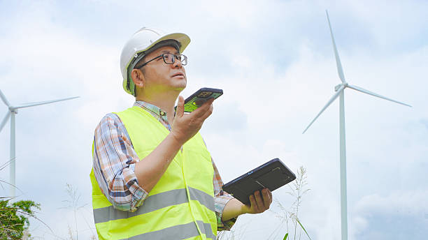 Engineer at a wind farm uses a smartphone and tablet simultaneously during a field inspection, coordinating maintenance tasks and monitoring windmill operations for clean, sustainable energy production.
