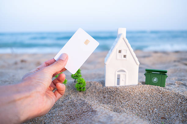 Holding a credit card near a miniature house and recycling bin on a sandy beach, suggesting themes of real estate investment and sustainable living