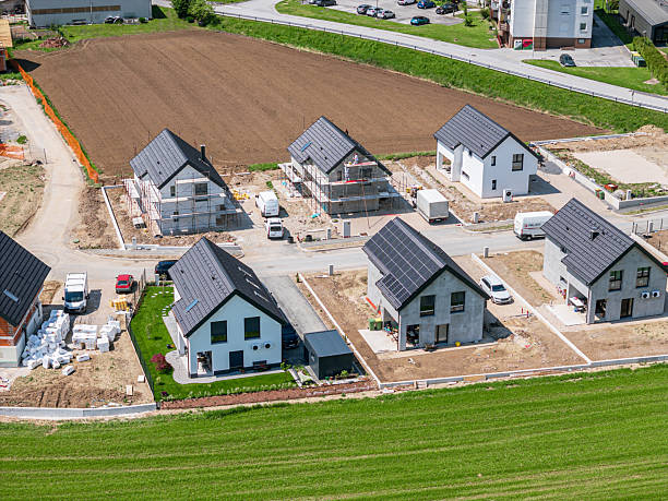 Aerial view of a residential construction site featuring several modern houses with solar panels. The area is partially landscaped with green grass and dirt paths, surrounded by fields and a road.