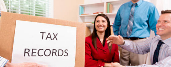 Cheerful, multi-ethnic firm of tax accountants work together on customer income tax returns. A client brings in a box of tax records in foreground. Two business men and one woman. Digital tablet, smart phones along with documents lie on desk.