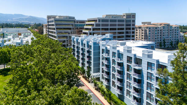 Aerial view of Cupertino's evolving cityscape showcasing a mix of modern architecture in California towns. Foreground features a sleek white residential complex with balconies and solar panels. Behind it, taller office buildings rise, their glass facades reflecting the California sun. Lush green trees line the streets, providing a natural contrast to the urban structures. The scene captures the essence of Silicon Valley's rapid growth and technological influence on urban planning.