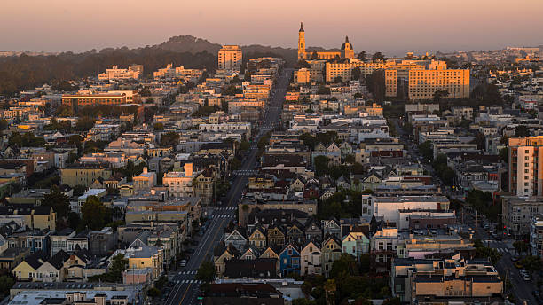 Aerial view of Mission District in San Francisco at sunrise with prominent church spires visible against pink morning sky. Straight road cuts through dense residential neighborhood with mix of Victorian houses and apartment buildings. Golden sunlight illuminates facades while Buena Vista Park creates dark silhouette in background. Historic architecture and urban planning of San Francisco visible from high angle view.