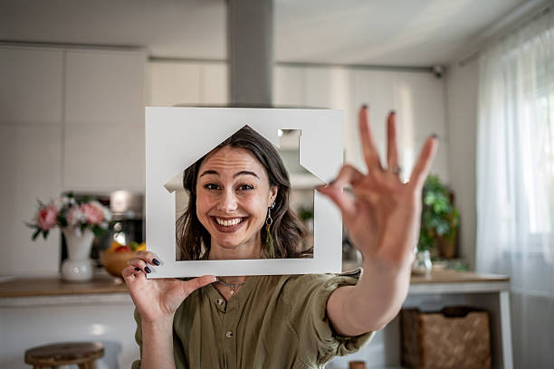 Happy young woman holding house shaped cardboard cutout in front of her face and showing ok sign with her hand, smiling and looking at camera in her kitchen