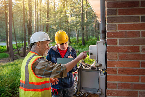 A 70-year-old supervisor in a white hard hat and safety vest demonstrates electrical inspection techniques to a 19-year-old apprentice with autism wearing a yellow hard hat. They examine an open electrical box mounted on a brick wall at a Pennsylvania worksite. The young man is focused on the task during his first job experience. Forest surrounds the work area with sunlight filtering through trees in the background. The senior worker points at components inside the control panel while the apprentice observes the training process closely.