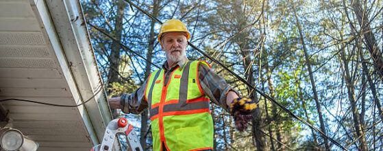 A senior construction worker in his 70s stands on a ladder while inspecting the roof of a residential building in Pennsylvania. He wears a bright yellow safety vest, blue jeans, and a yellow hardhat while holding inspection equipment. The worker is positioned near the eaves of the house with tall trees in the background. The scene shows routine home inspection or maintenance work during daytime with natural lighting filtering through the trees.