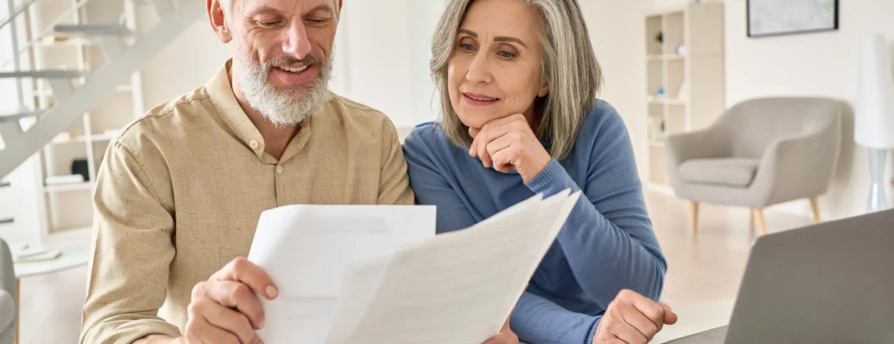 Middle aged senior old couple holding documents reading paper bills paying bank loan online, calculating pension fees, payments, taxes, planning family retirement money finances using laptop at home.