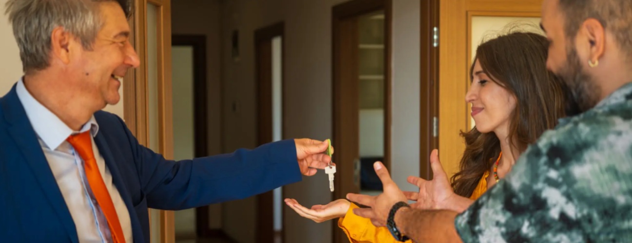 A smiling real estate agent hands over the house keys to a young couple inside their new home. The joyful expressions capture a milestone moment of ownership, trust, and achievement in the home-buying journey.