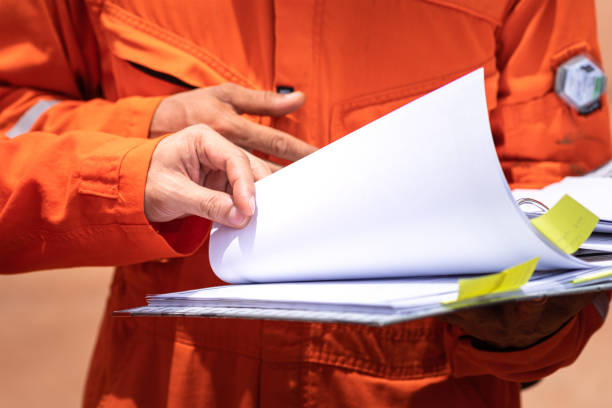 A worker in orange coverall uniform is opening documents to make a discussion on working procedure, safety audit in operation action photo. Close-up and partial focus on human's hand part.
