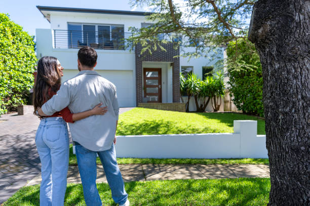 Happy Couple standing in front of their new home. They are both wearing casual clothes and embracing. Rear view. The house is contemporary with a brick facade, driveway, balcony and a green lawn. The front door is also visible.