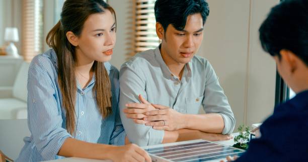 Closeup of young Asian couple in detailed consultation with real estate agent, who is highlighting advantages of home with solar panels. Consulting on Eco-Friendly Home with Real Estate Agent Concept.