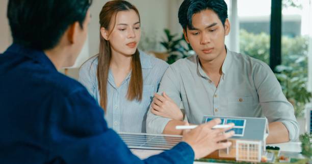 Young Asian couple in detailed consultation with real estate agent, who is highlighting advantages of home with solar panels. Consulting on Eco-Friendly Home Ownership with Real Estate Agent Concept.