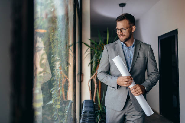 A confident businessman dressed in a gray suit, wearing glasses and a watch, stands indoors holding rolled up documents with a friendly and approachable smile. Portrait of multiethnic architect with blueprints in creative office. The background features indoor plants and modern lighting.
