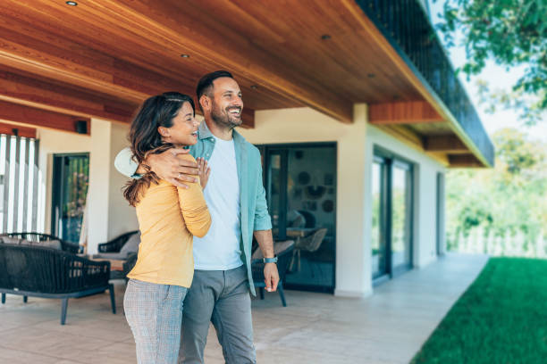 Couple standing in front of their new home. They are both wearing casual clothes and embracing. They are looking away and smiling. The house is contemporary with porchway and a green lawn, wood and glass exterior design