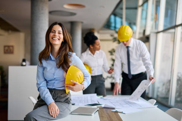 Portrait of a smiling female architect holding a yellow helmet in the office while her colleagues are working on blueprints in the background