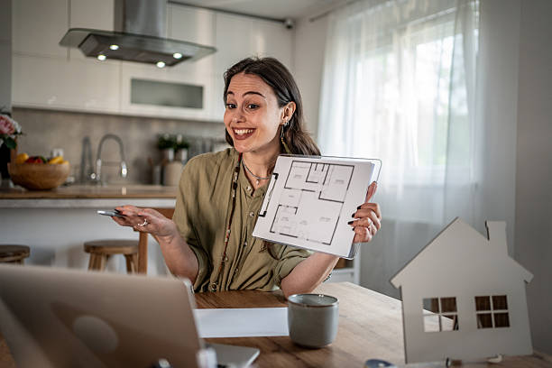 Young architect presenting a detailed house plan during a video conference call, utilizing her laptop and phone while working from the comfort of her home office
