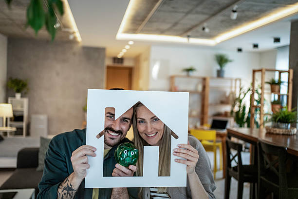 Smiling couple holding a house cutout and a green piggy bank, indicating their financial planning and aspirations for buying property and investing in their future home