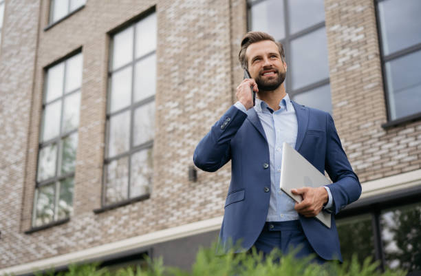 Handsome smiling businessman wearing stylish suit talking on mobile phone, holding laptop standing on the street. Successful business