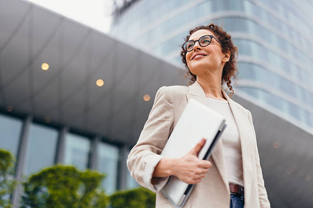 Happy businesswoman in eyeglasses holding a laptop, smiling confidently while looking away, standing near a modern office building on a bustling city street
