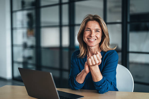 Confident and friendly professional mature woman sitting at a desk in a modern office setting. Senior Businesswoman is smiling warmly, creating a welcoming and approachable atmosphere. She is wearing a deep blue blouse and has light brown, shoulder-length hair. A laptop is open on the desk in front of her, indicating she may be engaged in remote work, a video call, or some form of digital task.
