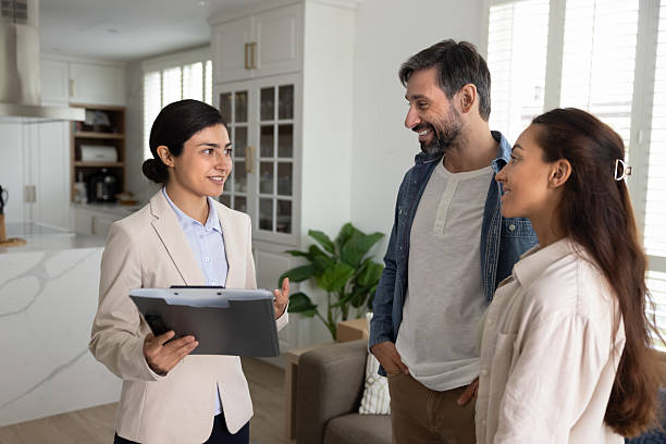 Agent explains details of agreement or investment documents to couple, standing together in modern house, talks about company services, conduct meeting with potential buying selling property clients