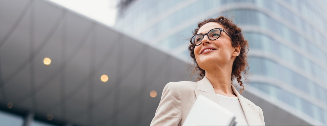 Happy businesswoman in eyeglasses holding a laptop, smiling confidently while looking away, standing near a modern office building on a bustling city street