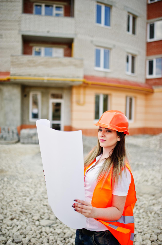 Engineer builder woman in uniform waistcoat and orange protective helmet hold business paper against new building property living block theme