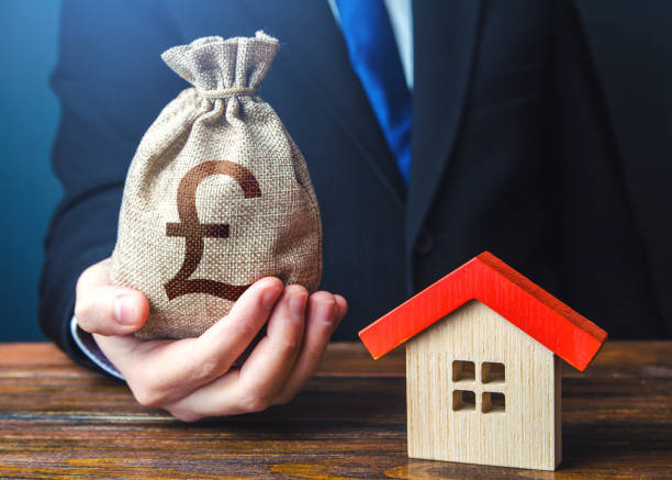 Man holds out a british pound sterling bag near the house. Home purchase, invest in real estate. Favorable terms and conditions, low interest rate. Bank approval for issuing a mortgage loan.