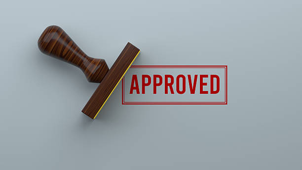 An official wooden rubber stamp has just made a bold, red "APPROVED" impression on a surface, viewed from directly above. This image is a clear and powerful symbol of success, validation, and positive confirmation. It represents the finality of a favorable decision, the achievement of a goal, or passing a quality control check. Ideal for business, finance, and legal applications, this visual effectively communicates concepts like loan approval, contract agreement, project completion, and certification, making it a perfect graphic for celebrating successful outcomes.