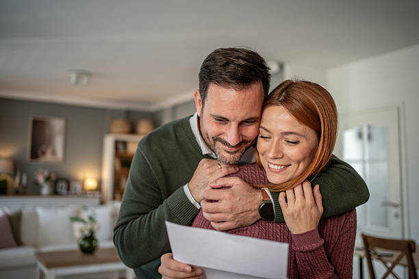 Loving couple happily embracing while reading an important letter with good news, sharing a tender moment of joy and financial success together in their comfortable home