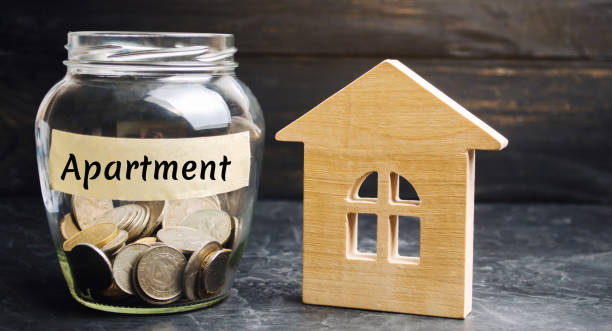 A glass jar with coins and a wooden house and the inscription "Apartment". The accumulation of money to buy an apartment. Rent an apartments, house. Rental housing. Saving money