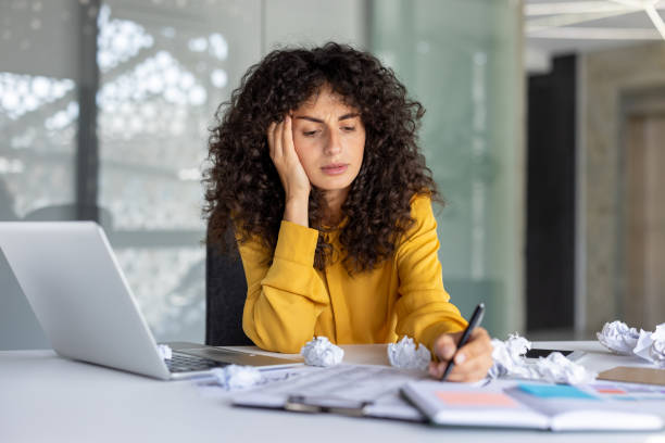 Frustrated woman at office desk, laptop open, surrounded by crumpled papers. Represents work stress, creativity block during busy day. Concept of workplace pressure and fatigue.
