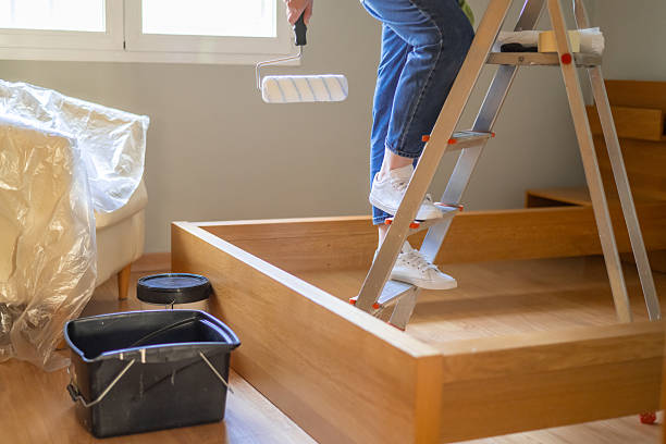 Person's legs standing on a metallic ladder holding a paint roller, preparing to paint a light grey wall in a bedroom during a home improvement project