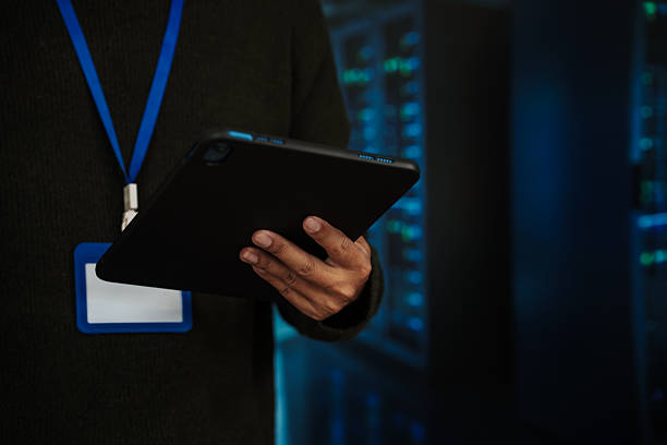 Close-up of it technician holding a tablet with id badge lanyard in a server room, blurred racks with blue lights in background, cybersecurity and network management concept.