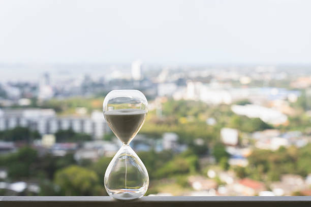 Hourglass resting on a window balcony with a blurred modern cityscape background. It represents time management, business deadlines, urban corporate investment, and future planning.