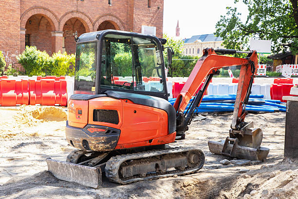 Compact crawler excavator at an urban roadworks zone. Professional heavy machinery operating on sand near red and white safety barriers. Compact crawler excavator at an urban roadworks zone. Professional heavy machinery operating on sand near red and white safety barriers.