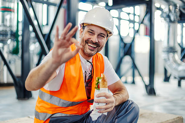 Mid adult construction worker wearing a hardhat and safety vest eating a sandwich and showing an ok gesture during his lunch break at an industrial facility, expressing job satisfaction